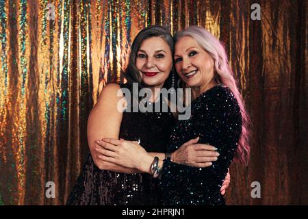 Two smiling senior girlfriends wearing evening dresses hugging each other while standing at glitter background Stock Photo