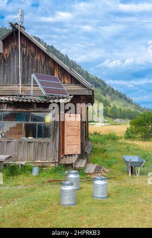 Old wooden house in mountains with solar panel on roof and milk cans on grass. The door is open. Wheelbarrow at the steps. Clouds in the sky. Vertical Stock Photo
