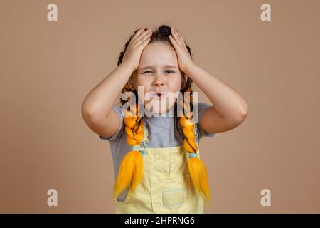 Regretting young child with yellow kanekalon pigtails, touching head with hands and with worried face in yellow jumpsuit and gray t-shirt on beige Stock Photo