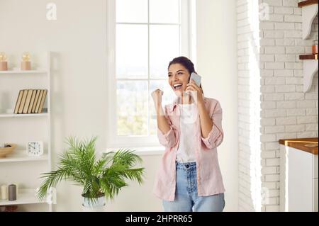 Joyful young woman sincerely rejoices hearing good news while talking on mobile phone at home. Stock Photo