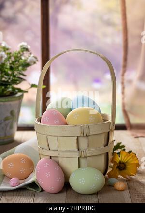 Colorful easter eggs in an old basket and on a rustic wooden table by a window with rural background Stock Photo