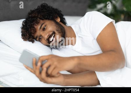 Smiling hindu guy laying in bed with smartphone Stock Photo