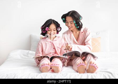 Glad happy millennial japanese lady and teen girl in pajamas and curlers doing pedicure and watching magazines Stock Photo