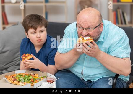 Dad and son are sitting on the sofa in the room eating pizza. Stock Photo
