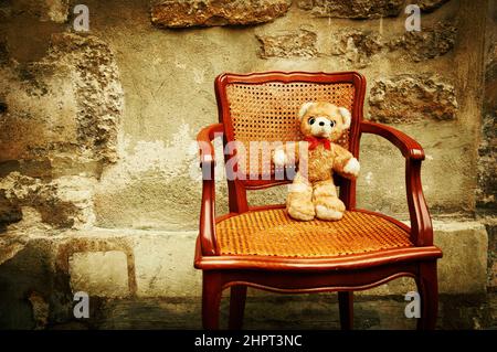 Teddy Bear sitting on chair and rough stone wall at background. Stock Photo