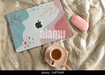 Macintosh laptop with colorful cover on a bed next to a mouse and a cup of tea. Working at home. Stock Photo