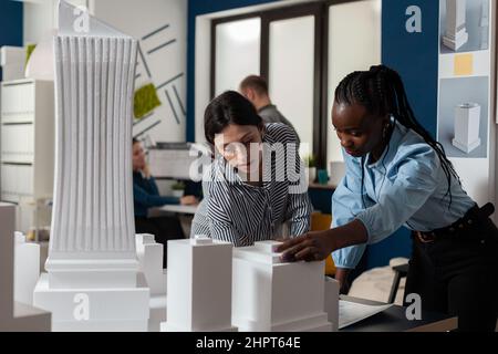 Architectural engineers doing teamwork reading construction plans over table with white foam scale model of buildings. Team of two architects colleagues collaborating looking down at blueprints. Stock Photo