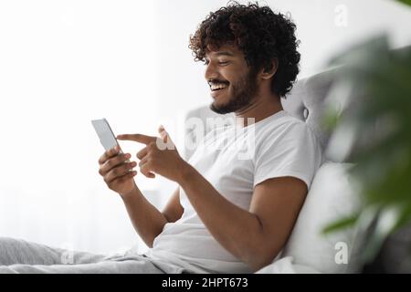 Positive dark-skinned guy scrolling in bed, using smartphone Stock Photo