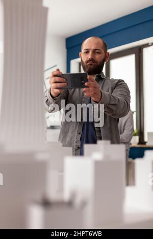 Architect using smartphone to record video of architectural maquette of skyscraper to post on social media. Bearded engineer photographing 3d white foam model with smart phone in modern office. Stock Photo
