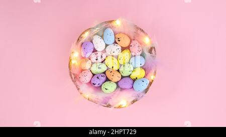 Creative wooden backet decorated with feathers and colorful eggs for Easter on pastel pink background. Minimal spring composition Stock Photo