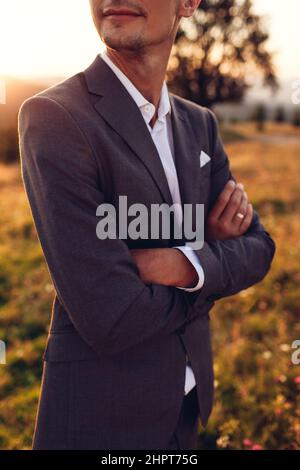 Handsome groom walking in wedding suit in mountains at sunset. Smiling man enjoys landscape with hands crossed Stock Photo