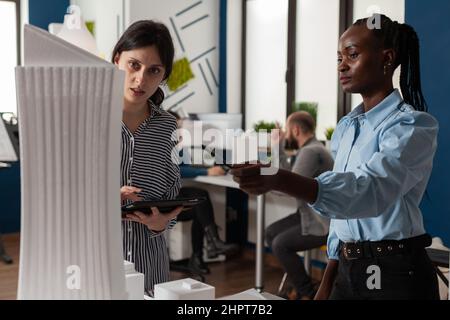African american woman architect pointing pencil at skyscraper architectural foam model standing next to colleague engineer holding tablet in modern office. Architects doing teamwork. Stock Photo