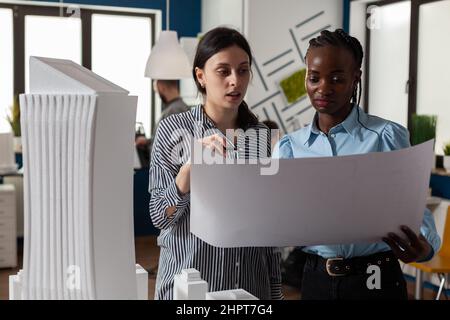 Team of two women architects reading architectural blueprints in front of desk with skyscraper 3d white foam scale model. Focused engineers doing teamwork holding construction plans. Stock Photo