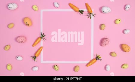 Pastel pink background with white copy space frama surrounded with Easter eggs and carrots. Spring holidays ornaments. Flat lay concept Stock Photo