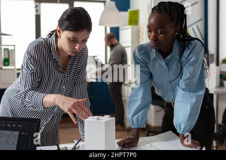 Architect colleagues discussing design of white foam building model in modern architectural office. Team of two professional architects working on blueprints on desk. Stock Photo