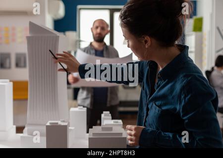 Architect inspecting futuristic building design of skyscraper with colleague engineer holding blueprints. Architects doing teamwork at desk with white foam maquette of construction project. Stock Photo