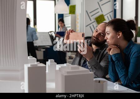 Architects doing teamwork looking at mobile content on smartphone sitting at desk in modern architectural office. Project engineers team collaboration using smart phone to photograph foam model. Stock Photo