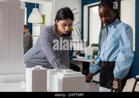 Architectural engineers working together reading construction plans standing next to table with foam model maquette. Team of two architects doing teamwork looking down at blueprins. Stock Photo