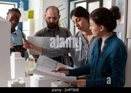 Two women architectural engineers looking down at blueprints next to colleagues holding laptop and tablet computer. Group of focused professional architects analyzing design plans. Stock Photo