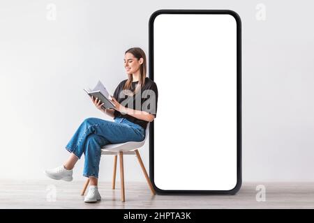 Young Caucasian woman reading book, sitting on chair near giant cellphone with empty screen, mockup. Cool mobile app Stock Photo