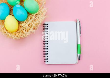 Blank notepad and pen on trendy dark pink background. Notebook for ideas message, list and inspiration. Easter eggs painted in pastel colors in hay. Stock Photo