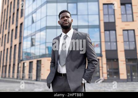 Young black man manager in suit holding hands in trousers pockets, standing against business center Stock Photo