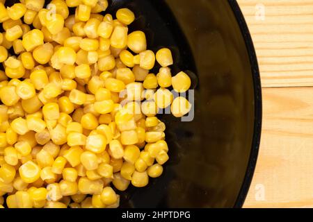 Bright yellow sweet kernels of canned corn in a black ceramic plate on a wooden table, macro, top view. Stock Photo