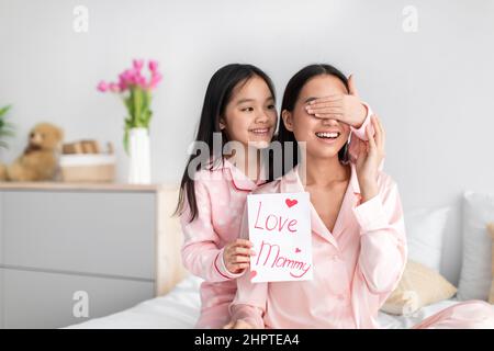 Smiling asian cute teen girl in pajama closes eyes to millennial woman hold postcard in bedroom interior Stock Photo