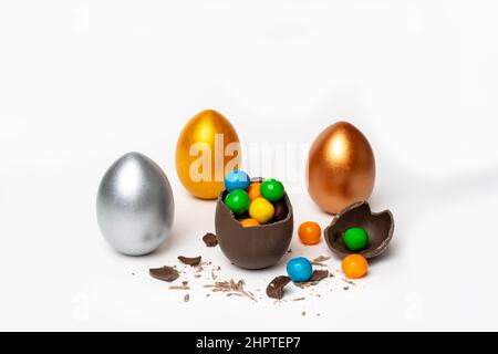 Cracked chocolate easter egg with colorful small round sweets and chocolates and whole colored eggs on white background, copy space. Easter concept. Stock Photo
