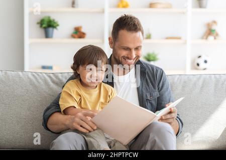 Storytime. Young loving father reading fairy tales to his cute little son, sitting with big child book on sofa at home Stock Photo