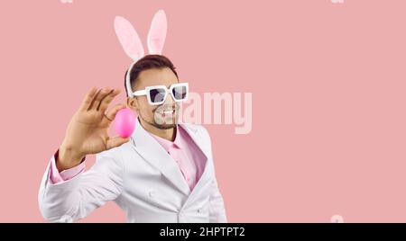 Handsome man in sunglasses and bunny ears showing Easter egg on pink copy space background Stock Photo