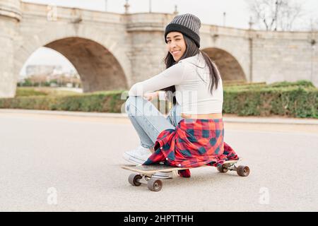 Portrait of a young woman with her back turned, wearing a cap, shirt tied at the waist and jeans, sitting on a skateboard in the middle of a road. Stock Photo