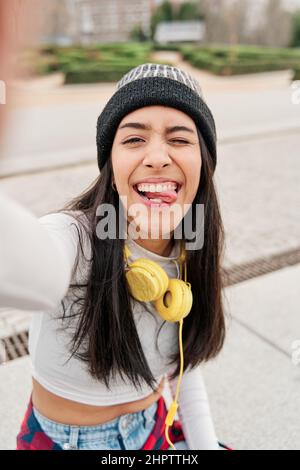 latina taking a selfie while making a funny face sticking out her tongue. Young Hispanic girl wearing casual clothes and a hat with headphones smiling Stock Photo