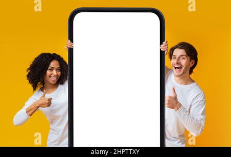 Cheerful interracial couple showing cellphone with blank screen, gesturing thumbs up, recommending mobile app, mockup Stock Photo