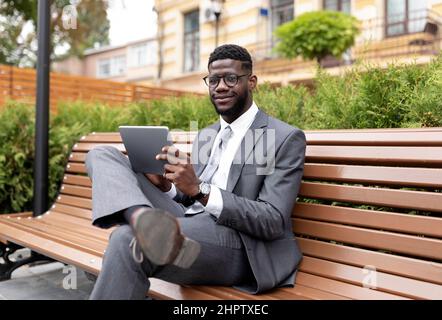 Confident african american entrepreneur using digital tablet, browsing news while sitting on the bench outdoors Stock Photo