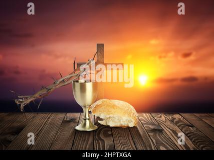 Chalice of wine, cross, bread and crown of thorns on table at sunset Stock Photo