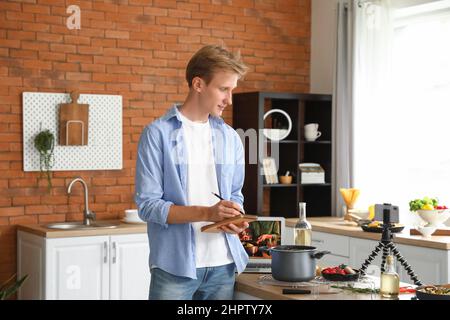 Young man making notes while watching video tutorial in kitchen Stock Photo