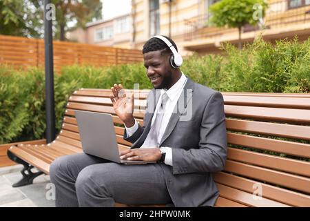Happy african american businessman having video conference, wearing headphones and waving hand to laptop webcamera Stock Photo