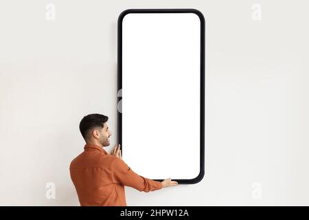 Young Arab guy demonstrating huge smartphone with blank screen, offering mockup for mobile app or website design Stock Photo