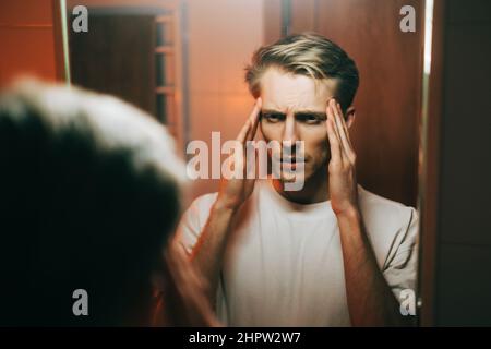 Young exhausted caucasian man standing in front the mirror and looking on himself, and touching the temples. Stock Photo