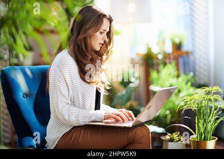 Green Home. stylish middle aged woman with long wavy hair at modern home in sunny day in beige pants and blouse using laptop. Stock Photo