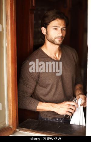 Long night ahead. Shot of a handsome young man drying off a glass while looking through a window. Stock Photo