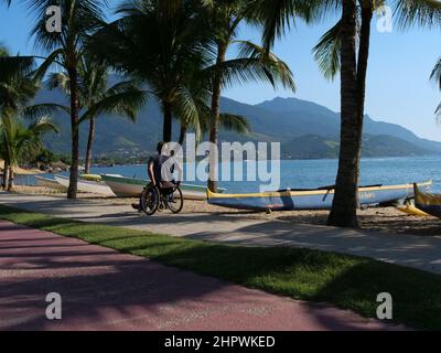 A disabled man in a wheelchair moves away from the viewer. He is using the pedestrian pathway on the island of Ilhabela, Brazil, Stock Photo