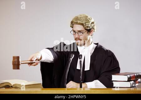 Silence. Serious young judge sitting in the courtroom with a stern facial expression while holding out a gavel. Stock Photo