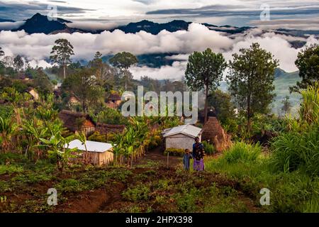 Woman and child outside of hamlet of thatched huts near garden, in PNG mountain valley, peaks in background, looking down on clouds. Incredible vista. Stock Photo