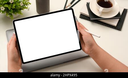 Female hand holding a digital portable tablet touchpad computer white empty screen mockup and stylus pen over office desk, closeup. Stock Photo