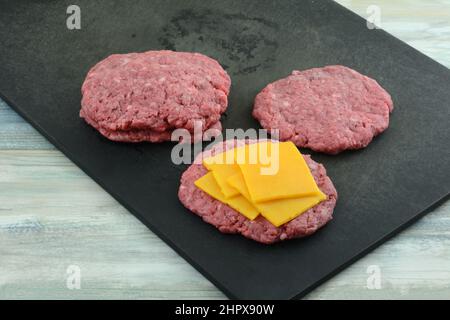 Placing cheese slices in between raw ground beef hamburger patties on black cutting board Stock Photo