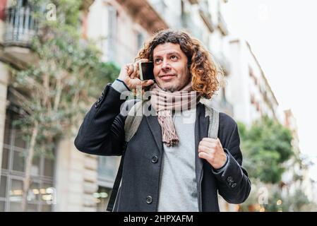 Low angle of positive mature male in smart casual clothes with backpack and scarf. He is looking away with smile and speaking on cellphone on blurred background of city street Stock Photo
