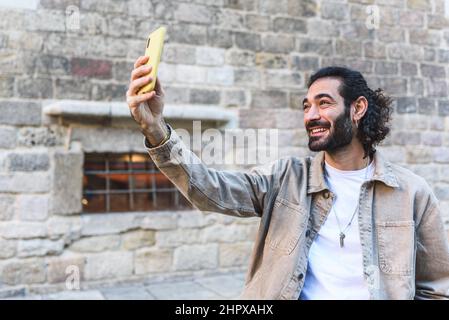 Delighted bearded male traveler smiling and taking self portrait on modern smartphone while standing near old building in city during trip Stock Photo