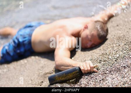 A drunk man with a bottle lies on the wet sand, blurry Stock Photo